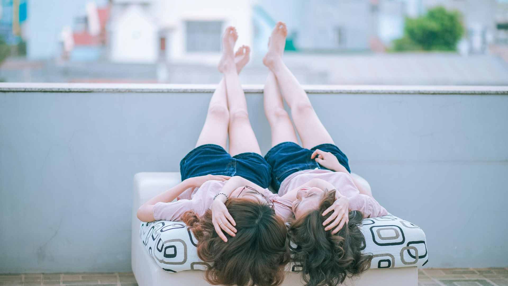 Two Woman Wearing Pink Shirt and Blue Shorts Lying on Sofa Facing Each Other While Both Feet Crossed and Placed on Concrete Wall