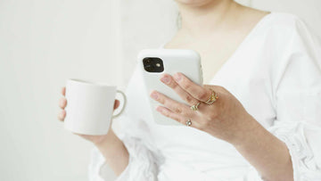  A Person in White Long Sleeves Holding a Cup of Coffee while Typing on Her Cellphone 