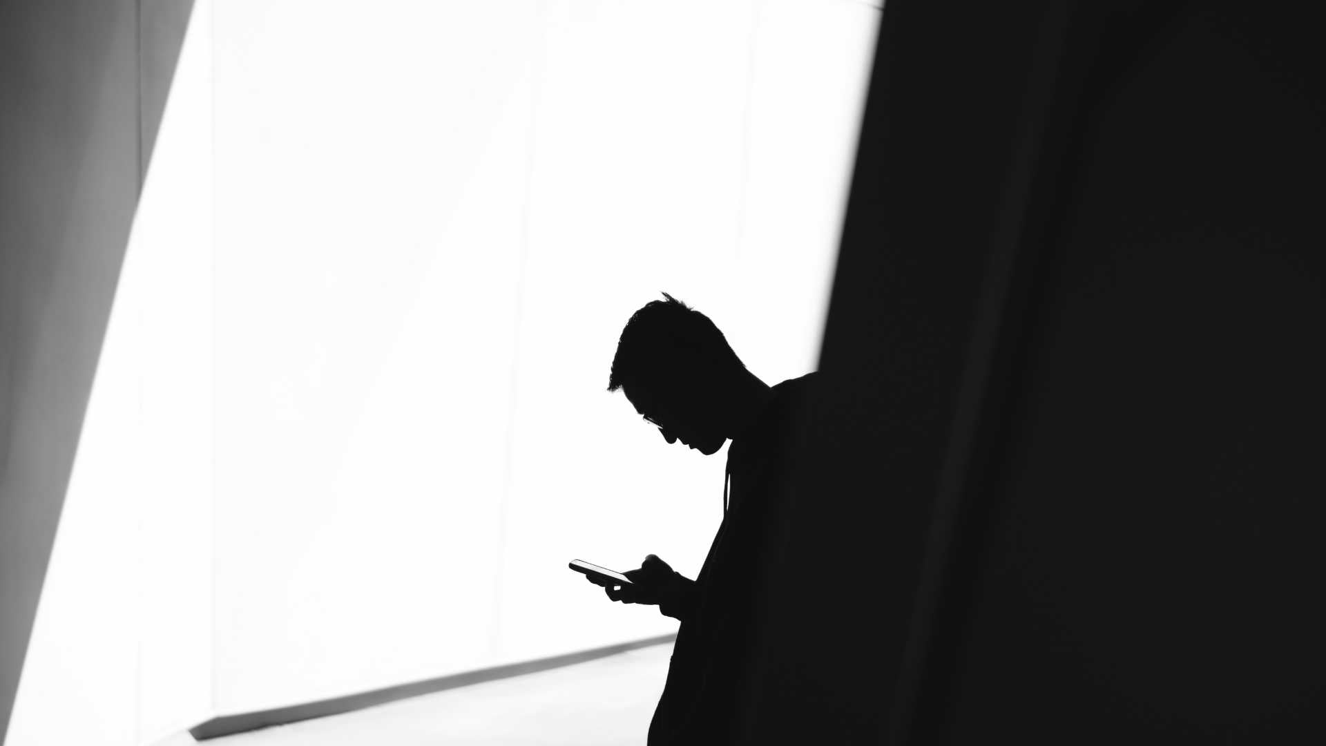 Person using phone leaning on wall in silhouette