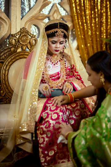 An Indian bride in rich traditional attire being helped with her outfit by a friend